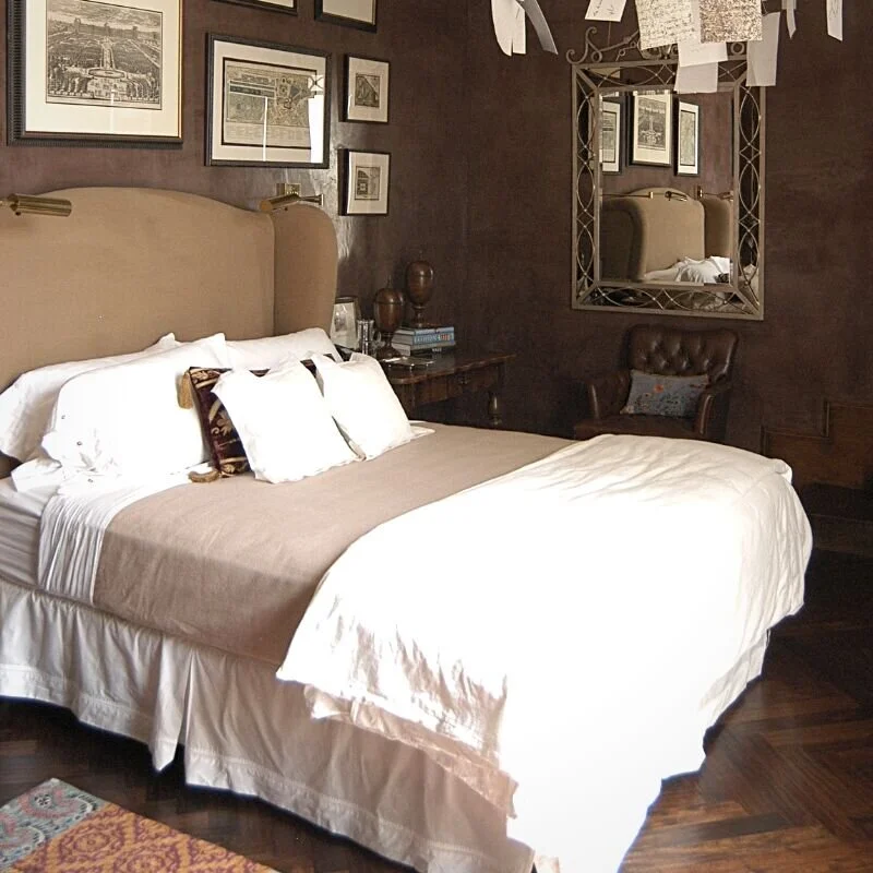 Photograph of a guest room with suede brown walls, with vintage drawings hanging.  The bed has an apolstered headboard and is made with a tan duvet and pillows.  There is a leather chair under a mirror in the corner. A room idea for when the kids move out.