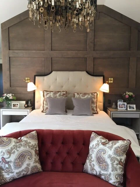 Bedroom with an upholstered headboard. The bed is flanked by reading lights and the wall behind the bed is geometric dark wood. In front of the bed is a red couch with throw cushions. A modern metal chandelier hangs above. This complements an article about choosing mattresses.