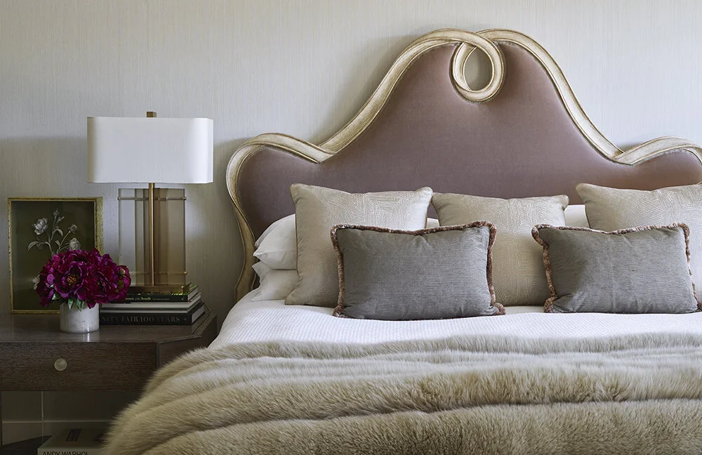 A close up of a bed and nightstand. The bed is appointed with a fuzzy blanket and throw pillows against an upholstered headboard. The side table has a modern rectangular lamp and a short vase of burgundy flowers.
