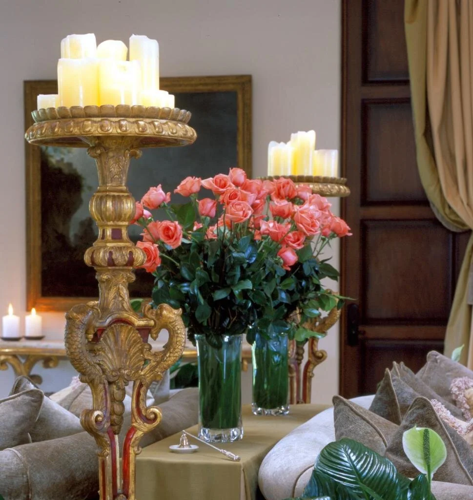 An ornate gold leaf candle stand as an investment piece. Behind is a sofa table with two vases of roses. The stand is topped with lit candles.